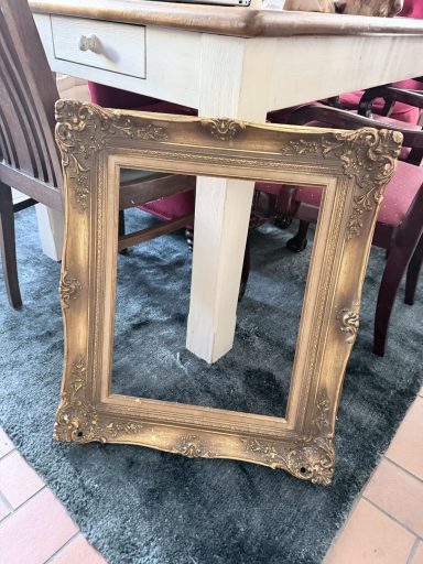 Barock-Rahmen Gold Ein vergoldeter Bilderrahmen steht auf einem Teppich neben einem Tisch und Stühlen.